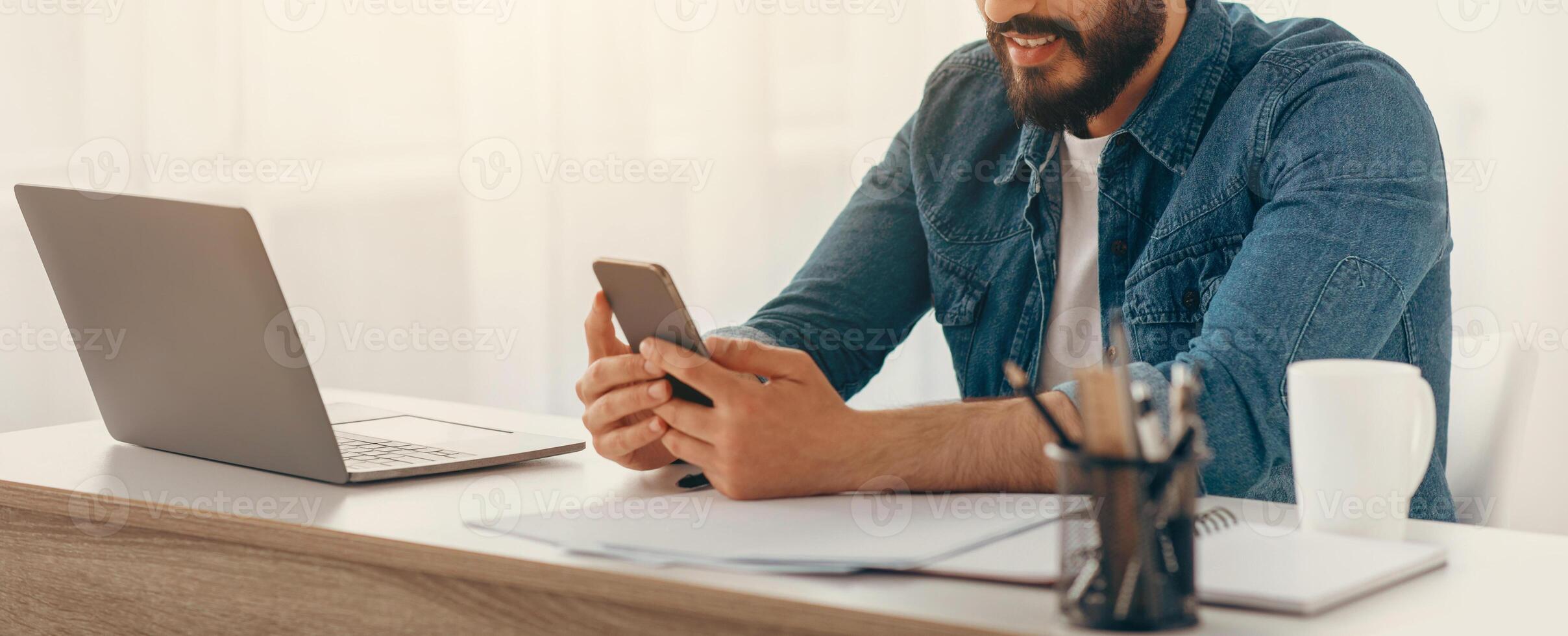 Arab freelancer guy using cellphone while working on laptop at desk at home office, browsing new app or messaging with friends, sitting at workplace with computer, free space photo