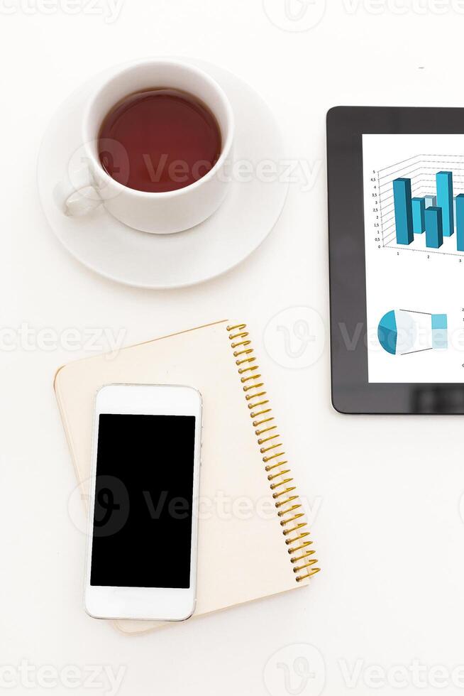 Busy workspace with coffee, smartphone, notebook, and tablet displaying data graphs photo