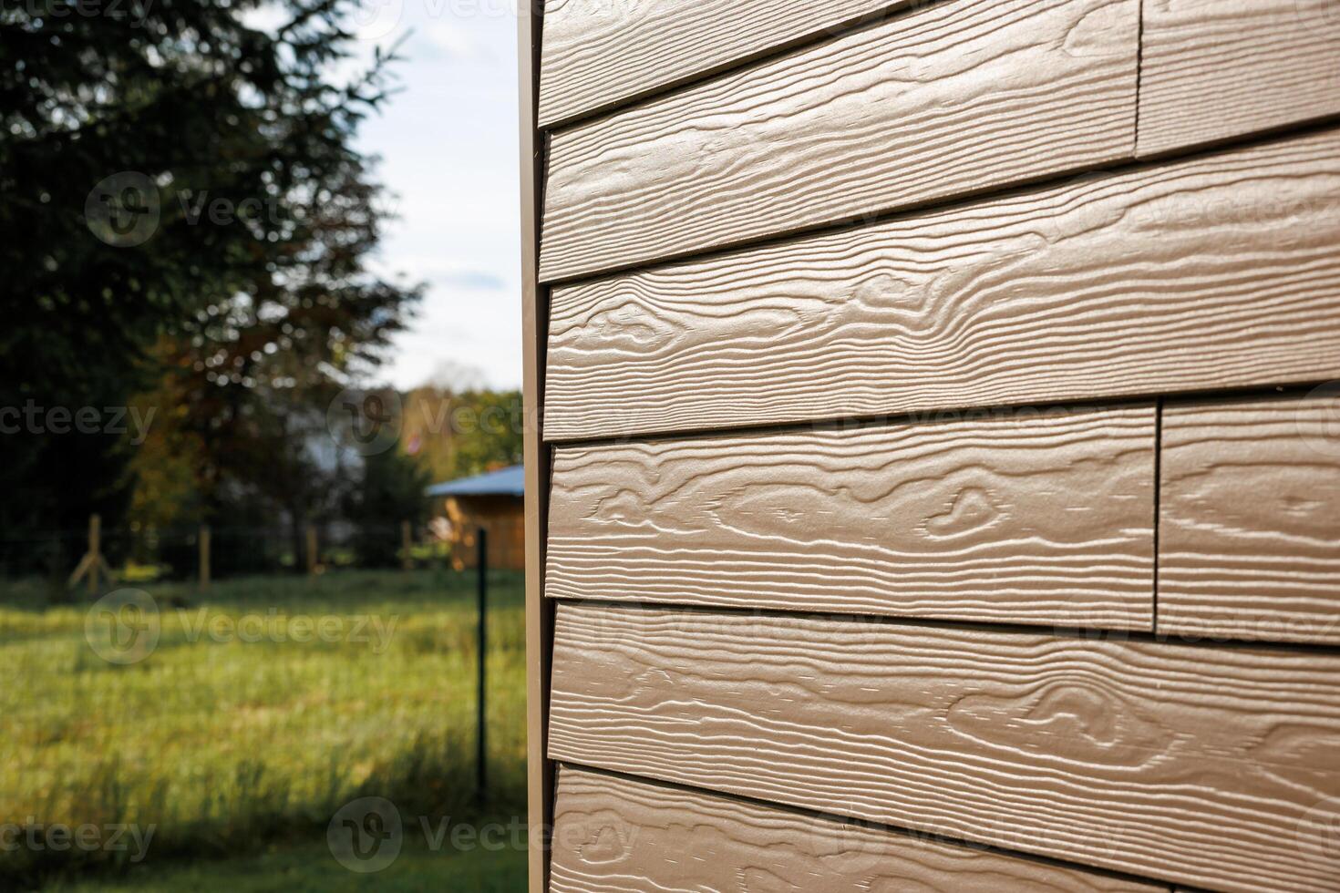 Textured beige horizontal siding with visible wood grain pattern, grassy area with fence, trees, and a small structure in the distance. photo