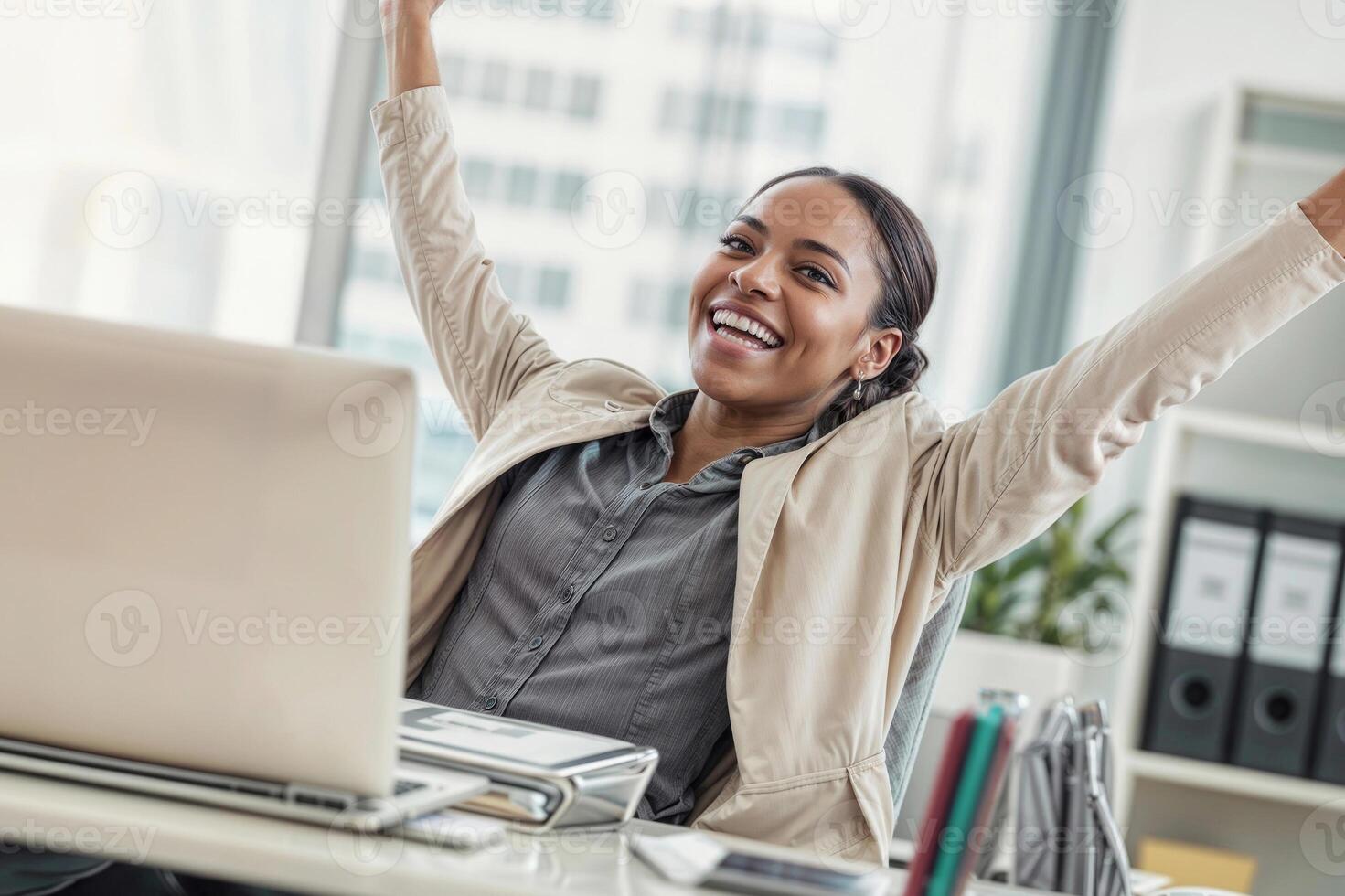 A woman is smiling and laughing while sitting at a desk with a computer, she is wearing a business suit and she is happy and excited rejoicing success photo