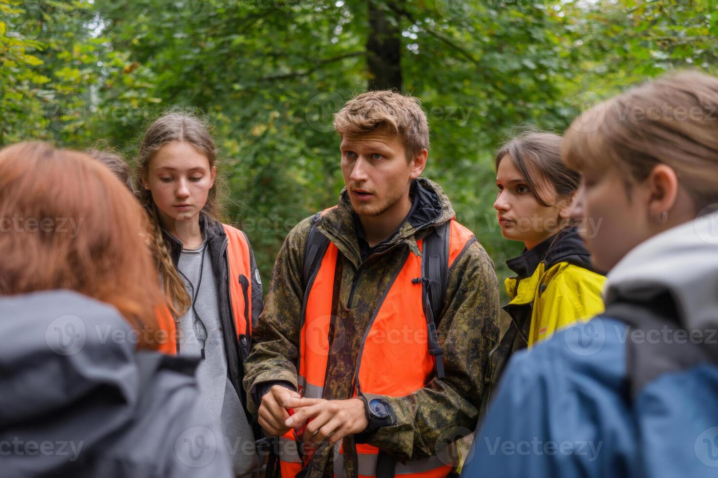 A group of young learners listens intently to a guide dressed in bright orange while exploring a green forest. They engage in a discussion about nature photo
