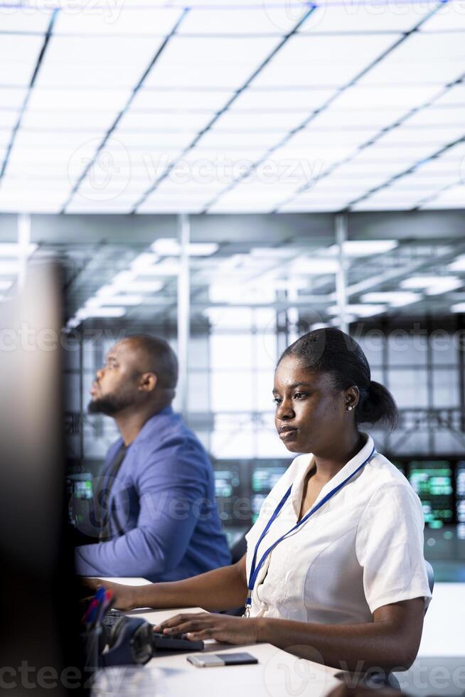 Technician and colleagues doing checkup to prolong data center electronics life span and avoid downtime. Woman in server hub monitoring server infrastructure components using computer photo