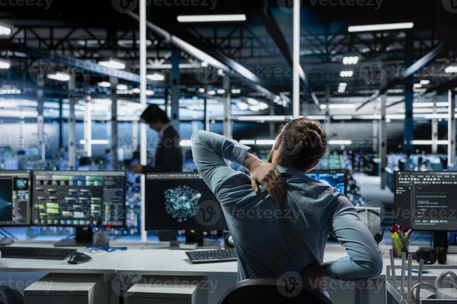 Tired data center software developer stretching to ease back tension while using AI LLM assistant. Server room IT professional massages injured back after long hours at computer coding with AI chatbot photo