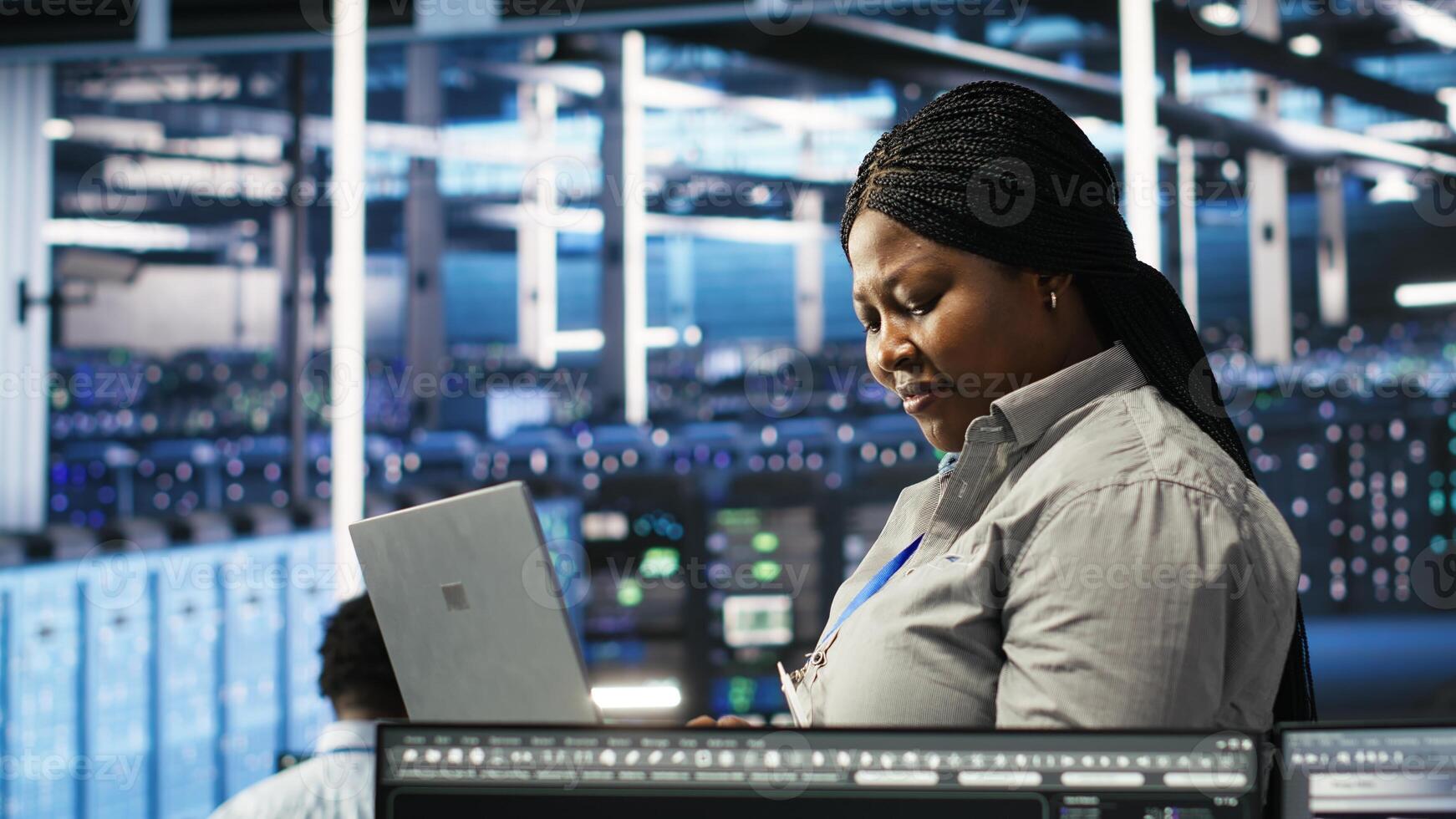 Data center admin using laptop to do node tree programming, optimizing resource allocation. Woman in server room uses predictive AI visualization systems on notebook for maintenance planning, camera B photo