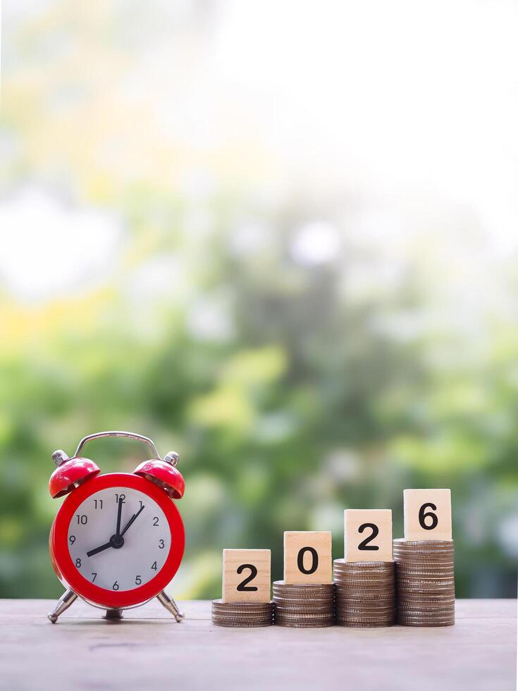 Red alarm clock, Wooden blocks with the number 2026 on stack of coins. Concept of saving money for manage time to success business in year 2026 photo