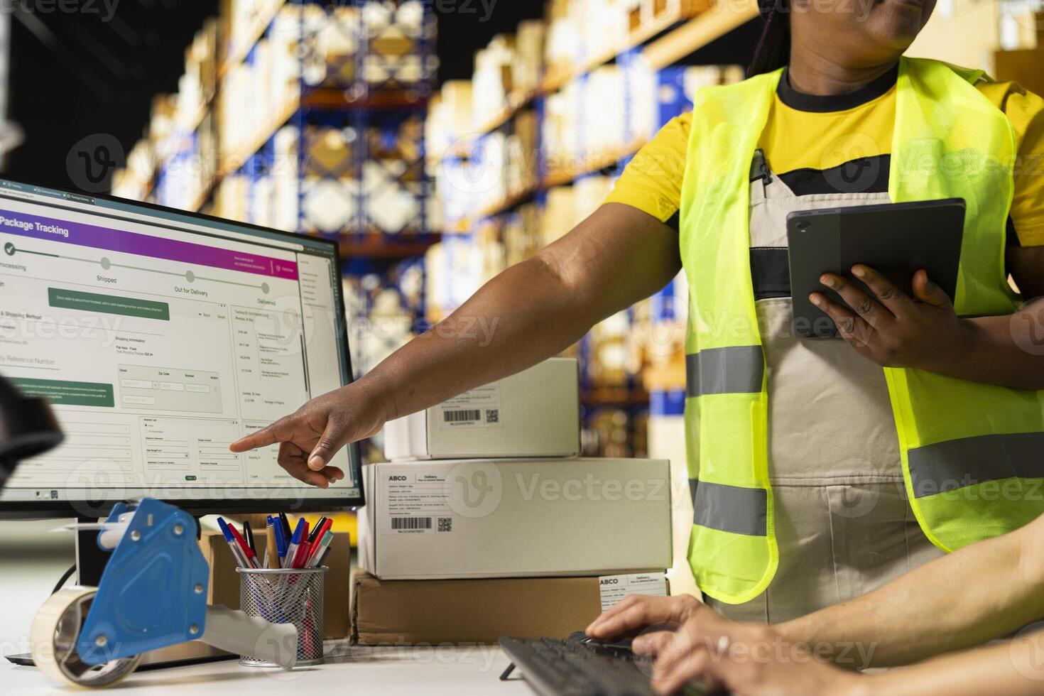 Black woman supervisor pointing at the parcel status on computer, using tracking software for packages shipment details. People work in depot filled with cardboard boxes on big racks. photo