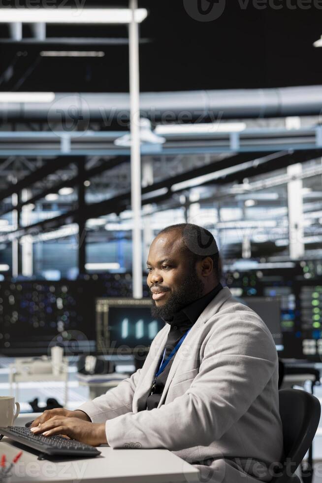 Upbeat data center IT expert uses PC to update servers tech, configuring gear using software. Jolly server room programmer reviewing hardware analytics, ensuring reliable network operations photo