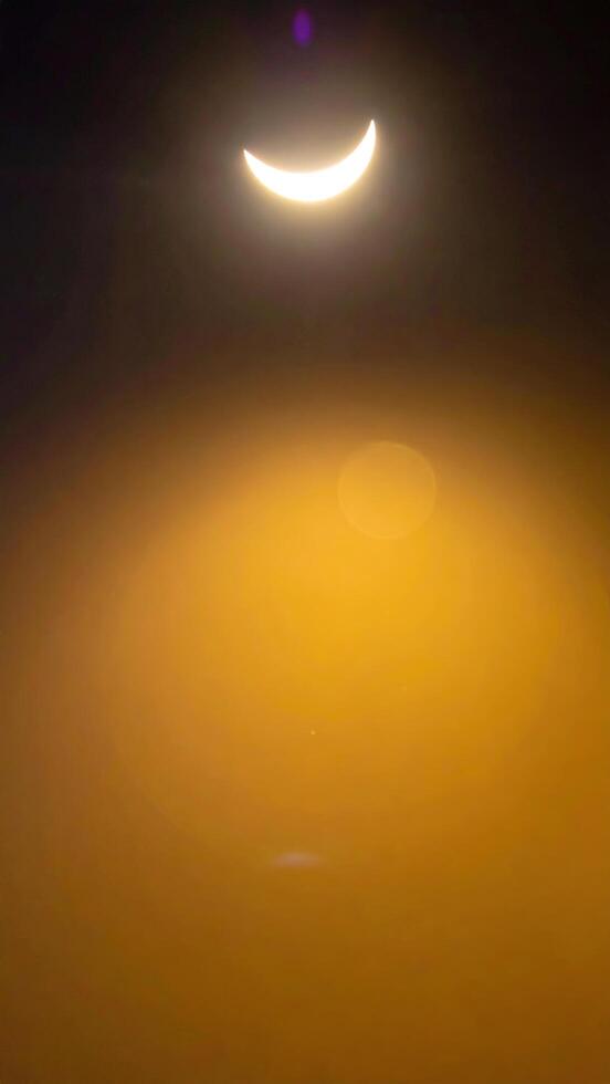 Celestial event captures the sun partially obscured by the moon, creating a crescent shape. Lens flare adds a vibrant golden glow and hazy atmosphere. An atmospheric halo is visible photo
