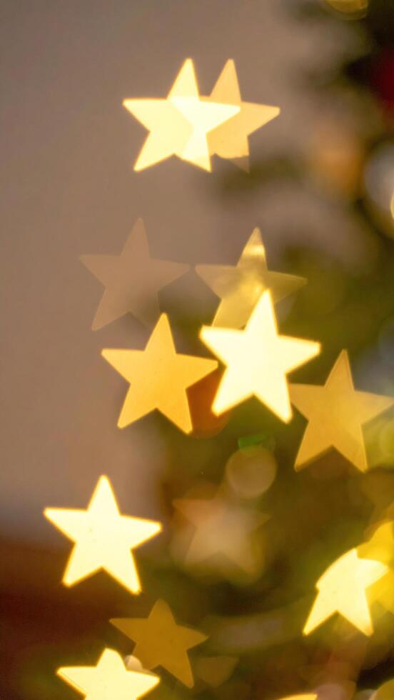 Soft-focused photograph featuring warm, yellow star-shaped lights, creating a bokeh effect. They partially obscure a Christmas tree's green branches, casting a festive glow against a neutral backdrop photo