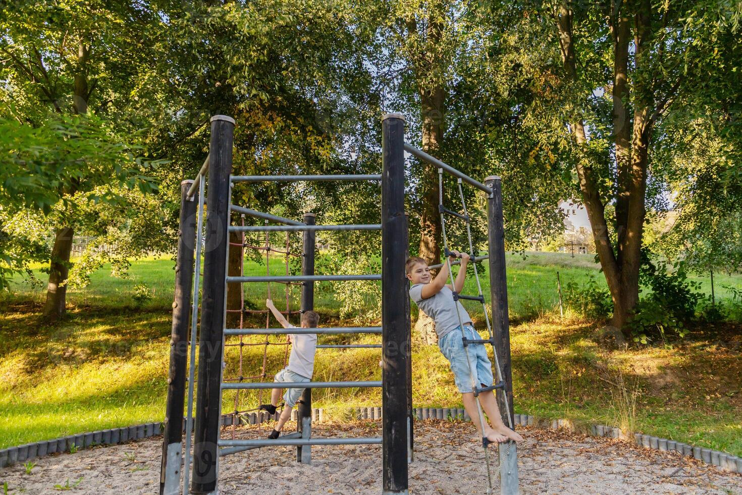 Two children are having fun playing on climbing bars in a nature playground. The warm sunlight filters through the trees, creating a joyful atmosphere in this outdoor setting photo
