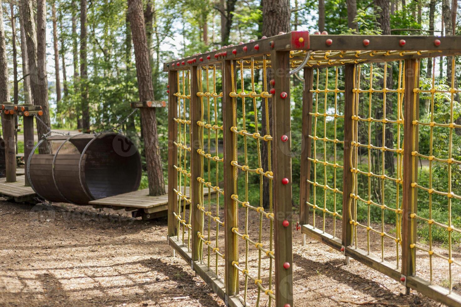 A climbing structure with colorful netting stands in a forest park, alongside a wooden barrel for children to play on. Lush greenery and trees create a cheerful atmosphere photo