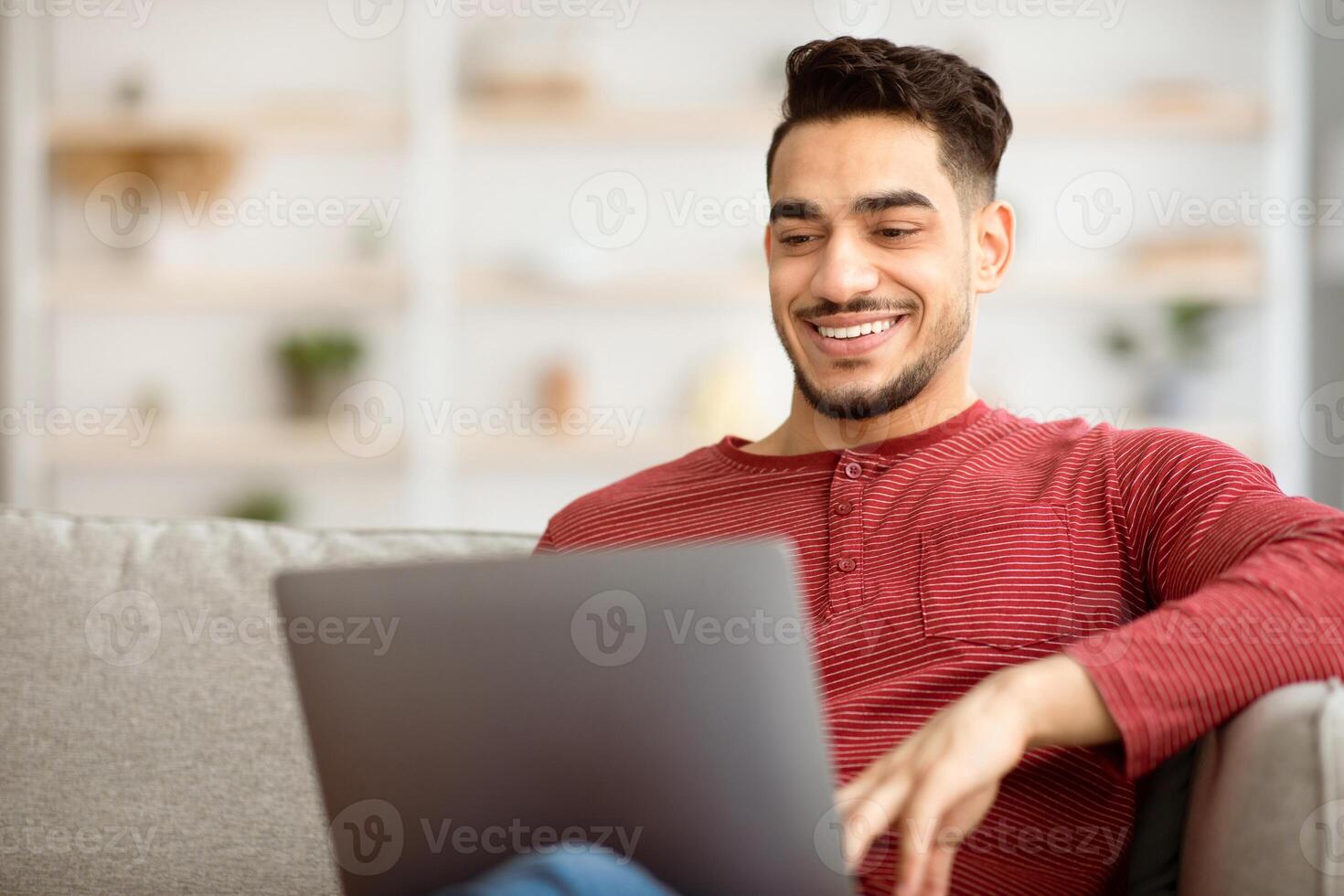 Closeup of happy middle-eastern man freelancer working from home, sitting on couch in living room and typing on notebook keyboard, chatting with clients or sending emails, copy space photo