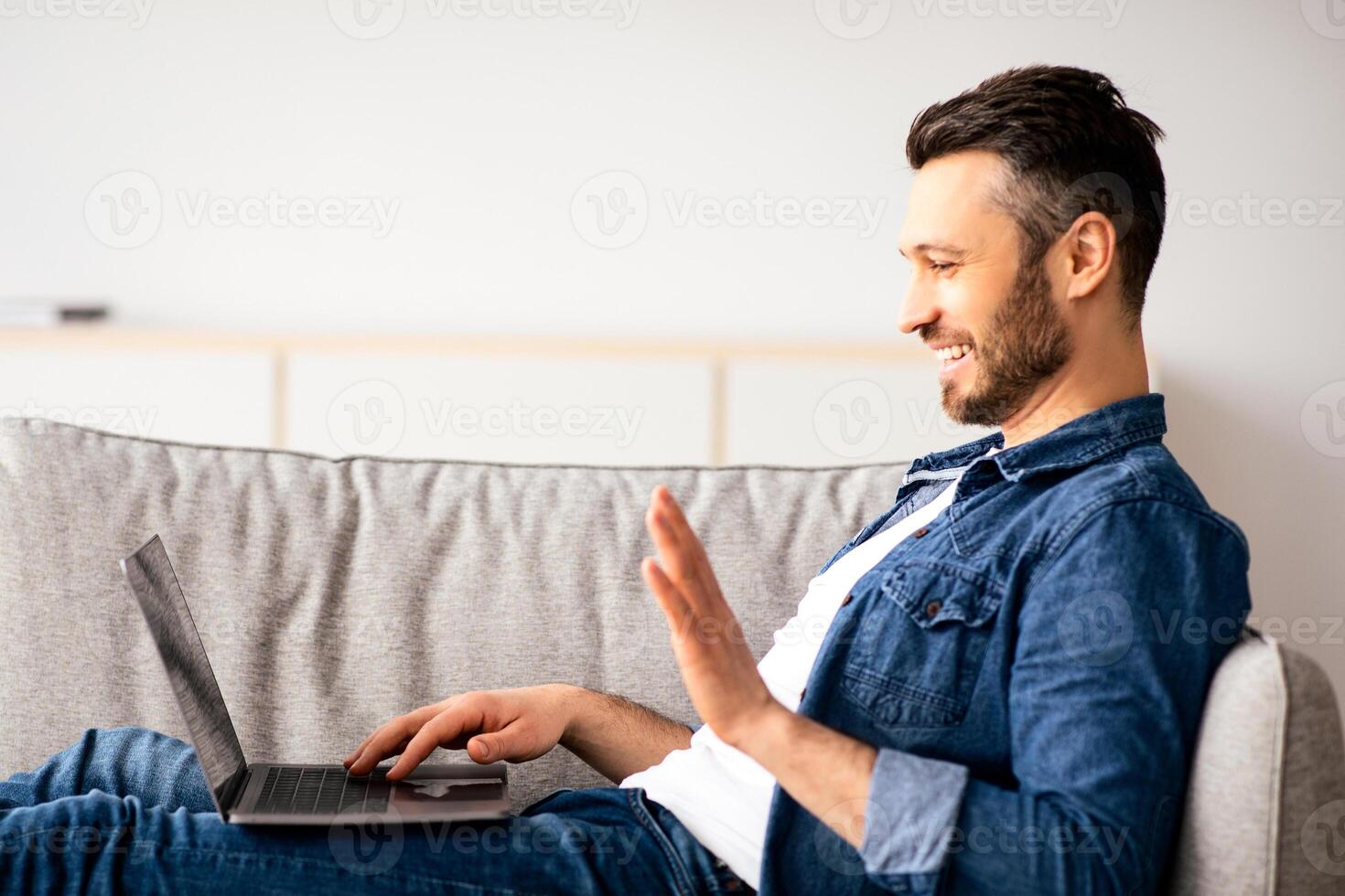 Smiling middle-aged bearded man having call with friends or lover, using laptop, looking at computer screen, talking and gesturing, home interior, copy space, side view photo