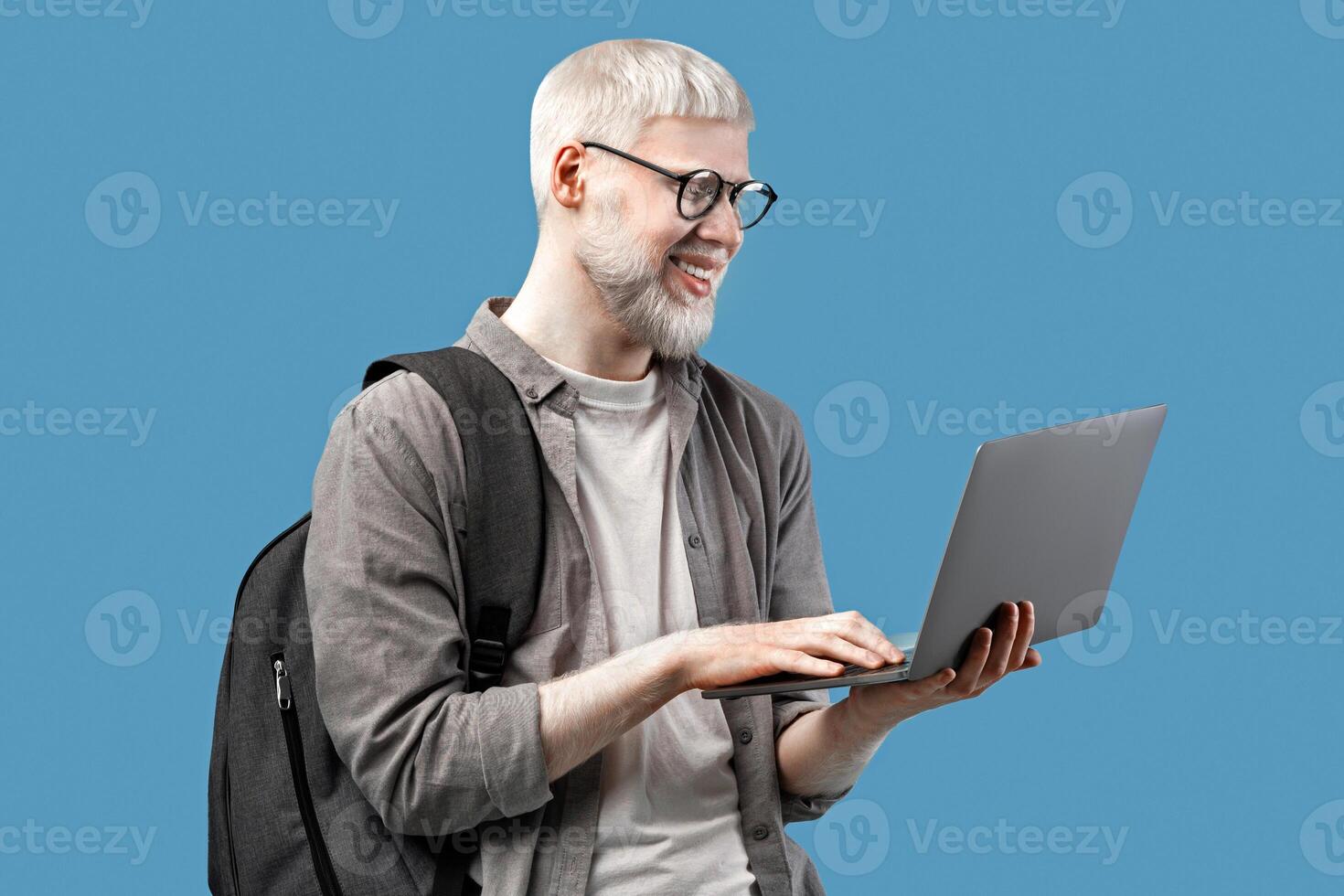 Cool guy with unusual appearance using laptop for online work or studies, wearing backpack over turquoise studio background. Happy young man communicating on web, browsing internet photo