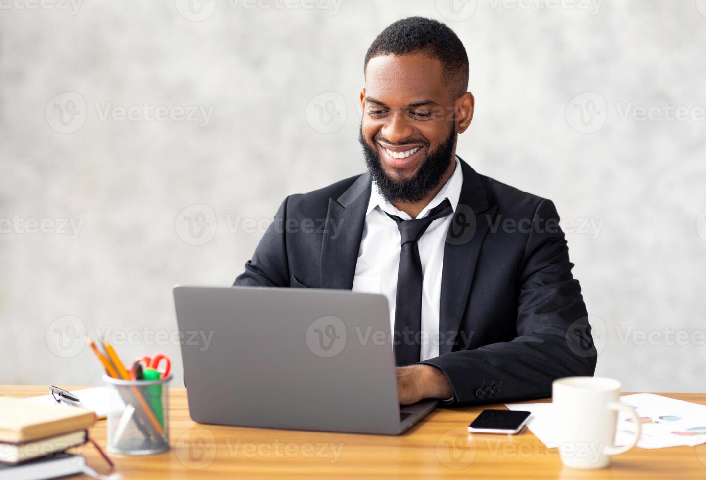 Portrait of handsome smiling African American male entrepreneur in suit working on laptop at desk in modern office, using computer at workplace, typing on keyboard, free copy space photo