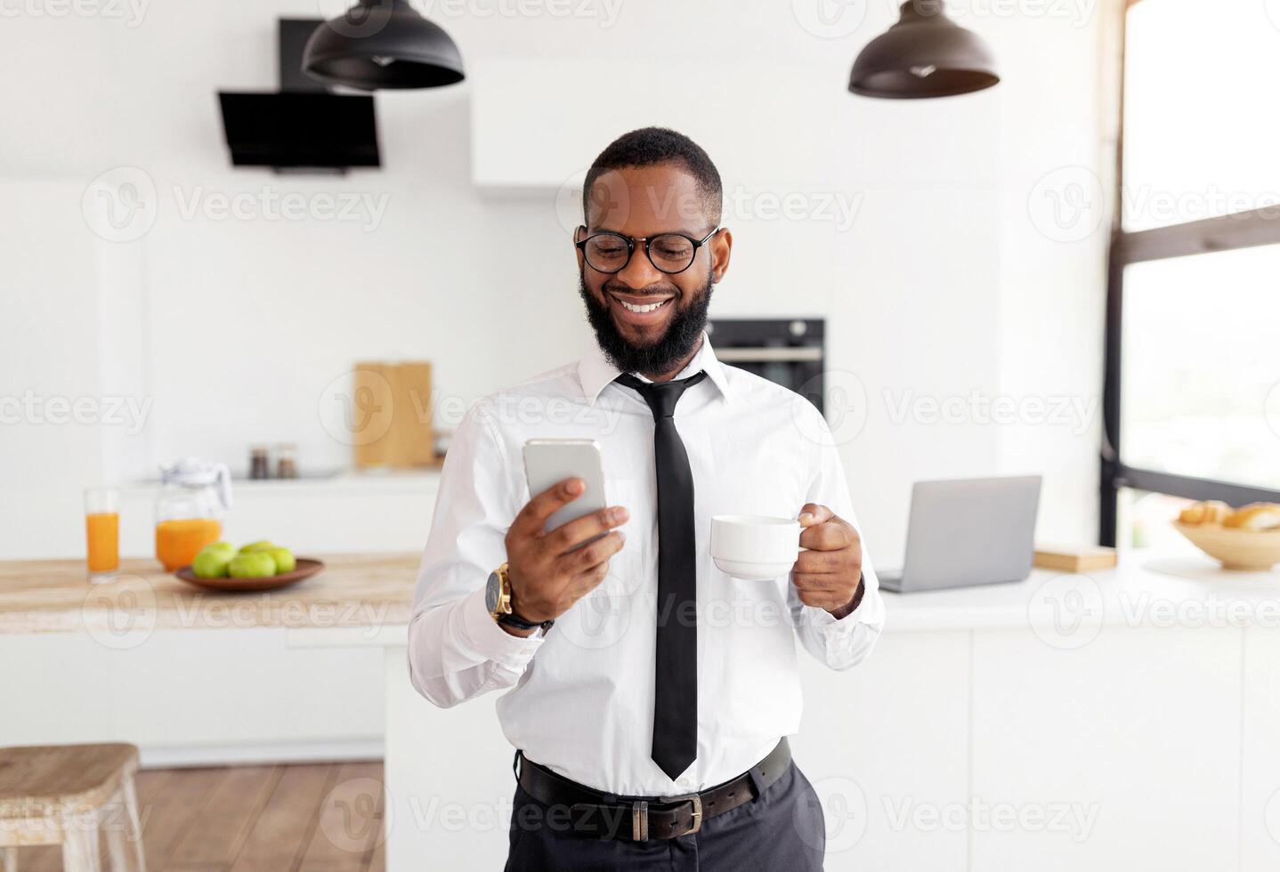 Smiling Black Business Man In White Shirt And Eyglasses Using Smartphone And Drinking Coffee While Standing At Home Office, Browsing Internet Or Social Networks, Enjoying Free Time And Break From Work photo