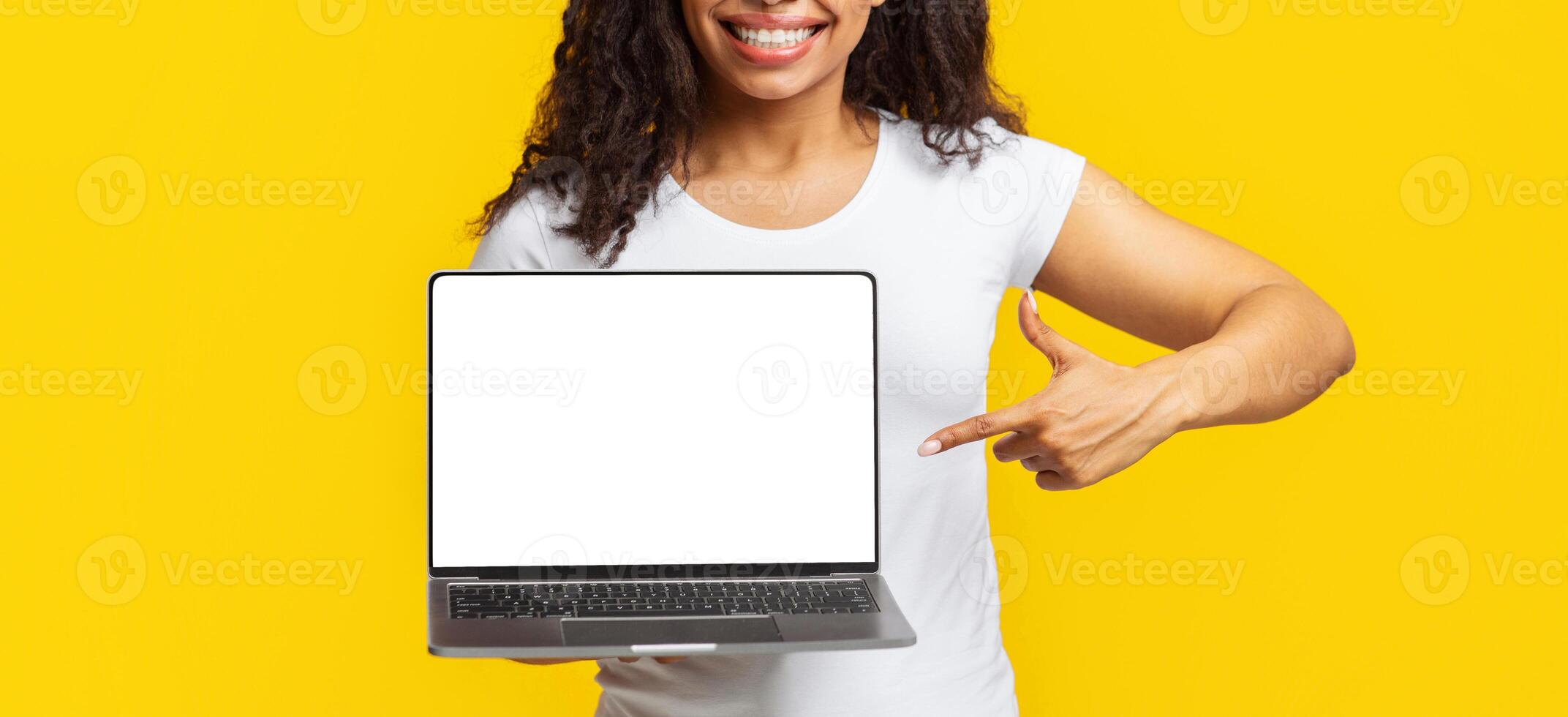 Online education. Happy black female student pointing at laptop with empty screen and smiling on yellow studio background. Positive lady with portable computer, mockup for design photo
