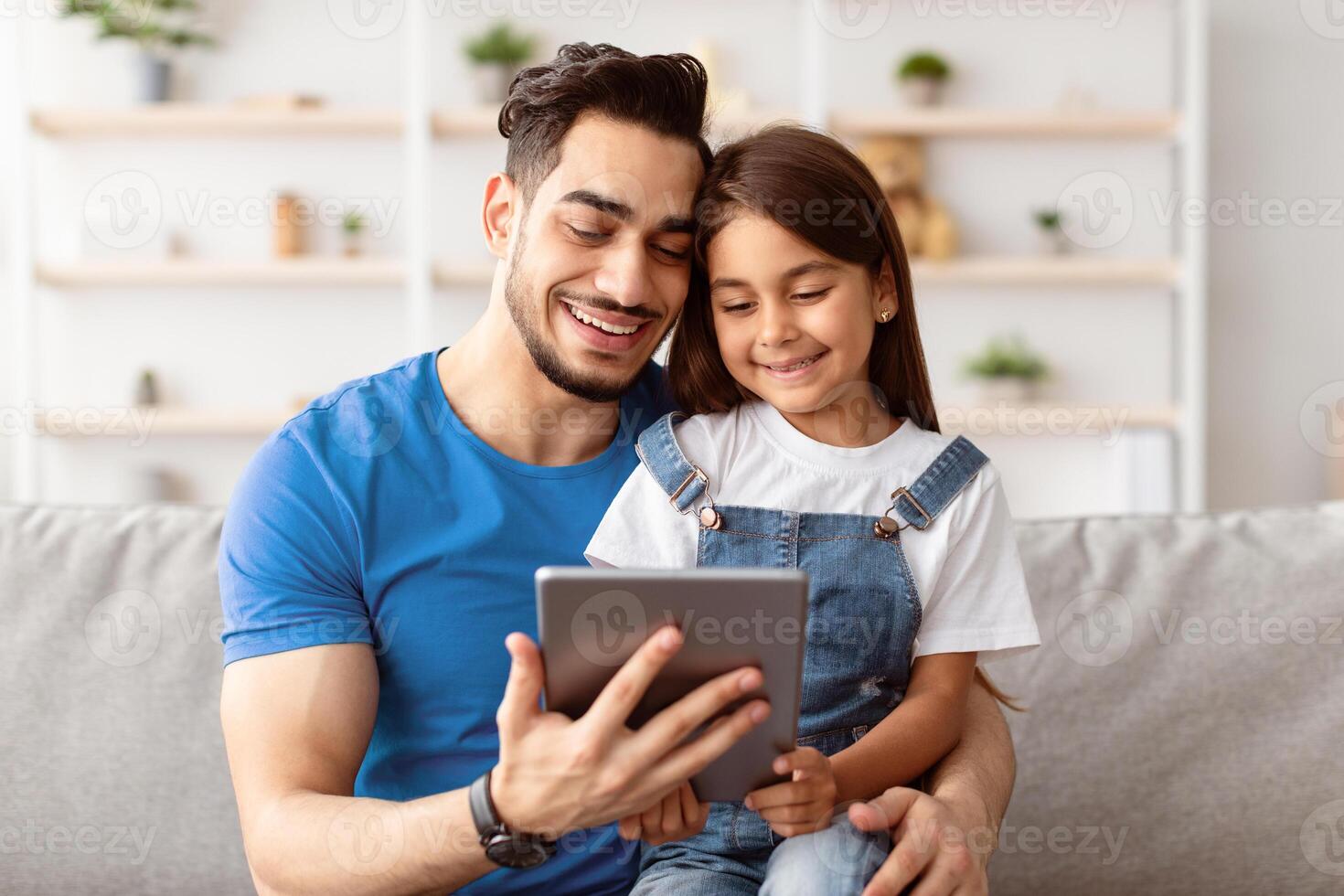 Young Father And Little Daughter Using Tablet Browsing Internet Together And Watching Movie Online Or Making Call, Sitting On Sofa At Home On Weekend. Daddy And Child Using Computer photo