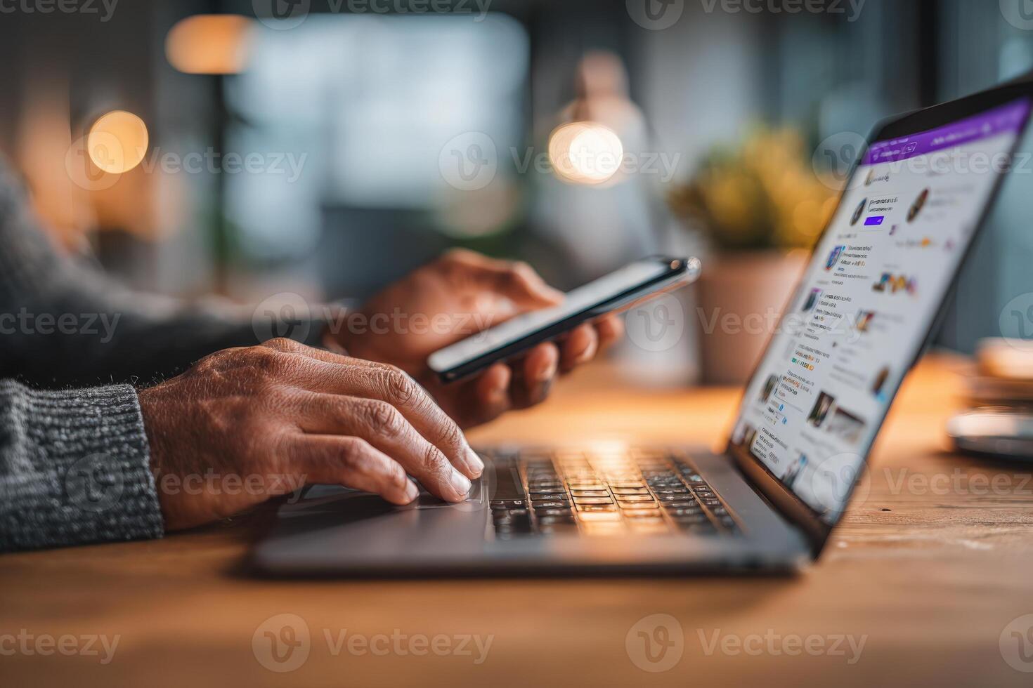 African American man using smartphone while typing on laptop, working in a modern office environment with warm lighting and wooden desk, showcasing multitasking and technology integration photo