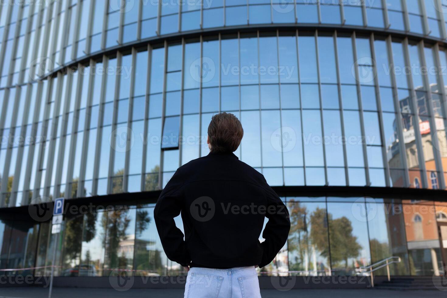 Man stands in front of large modern building reflecting blue sky and surrounding trees at midday photo