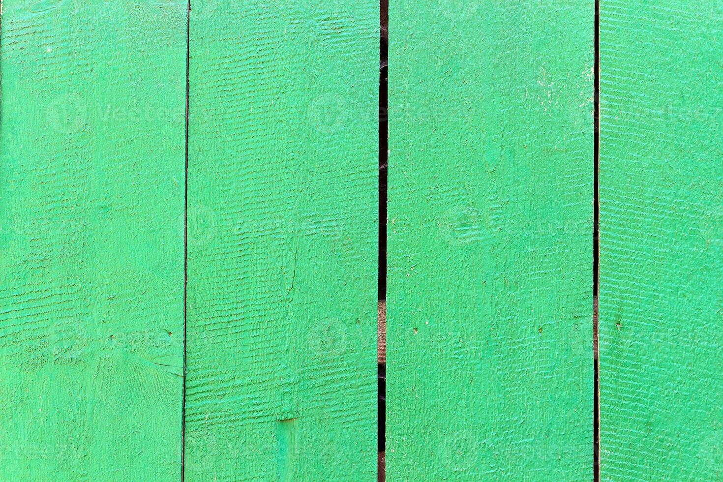 Green painted wooden fence with visible vertical planks and natural wood texture. Surface shows rough grain patterns, small cracks, and gaps between boards, creating rustic outdoor background photo