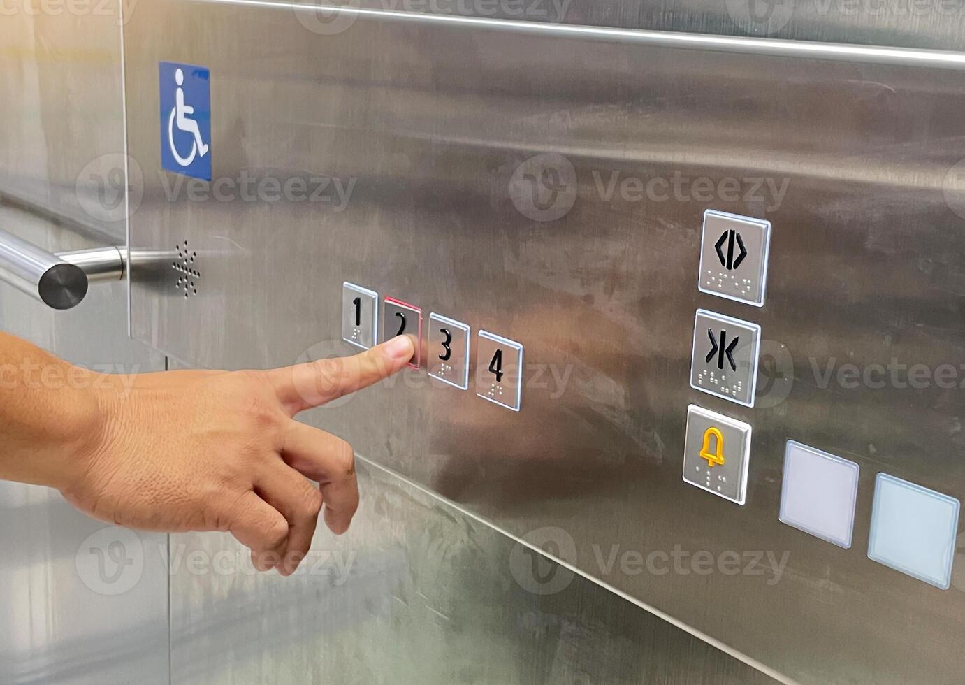 A hand presses the 2 button on a shiny metal elevator panel. The panel features floor numbers with Braille, a disabled access symbol, and other control buttons like door open-close and alarm. photo