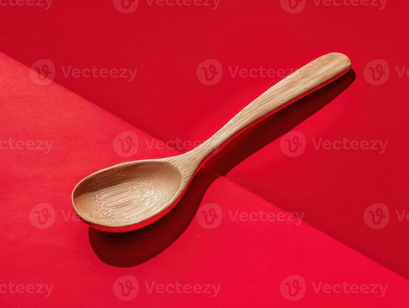 Wooden spoon rests diagonally on a smooth red surface, casting a shadow. The spoon's grain pattern is visible, and it appears to be made of a light-colored wood. No text is present photo