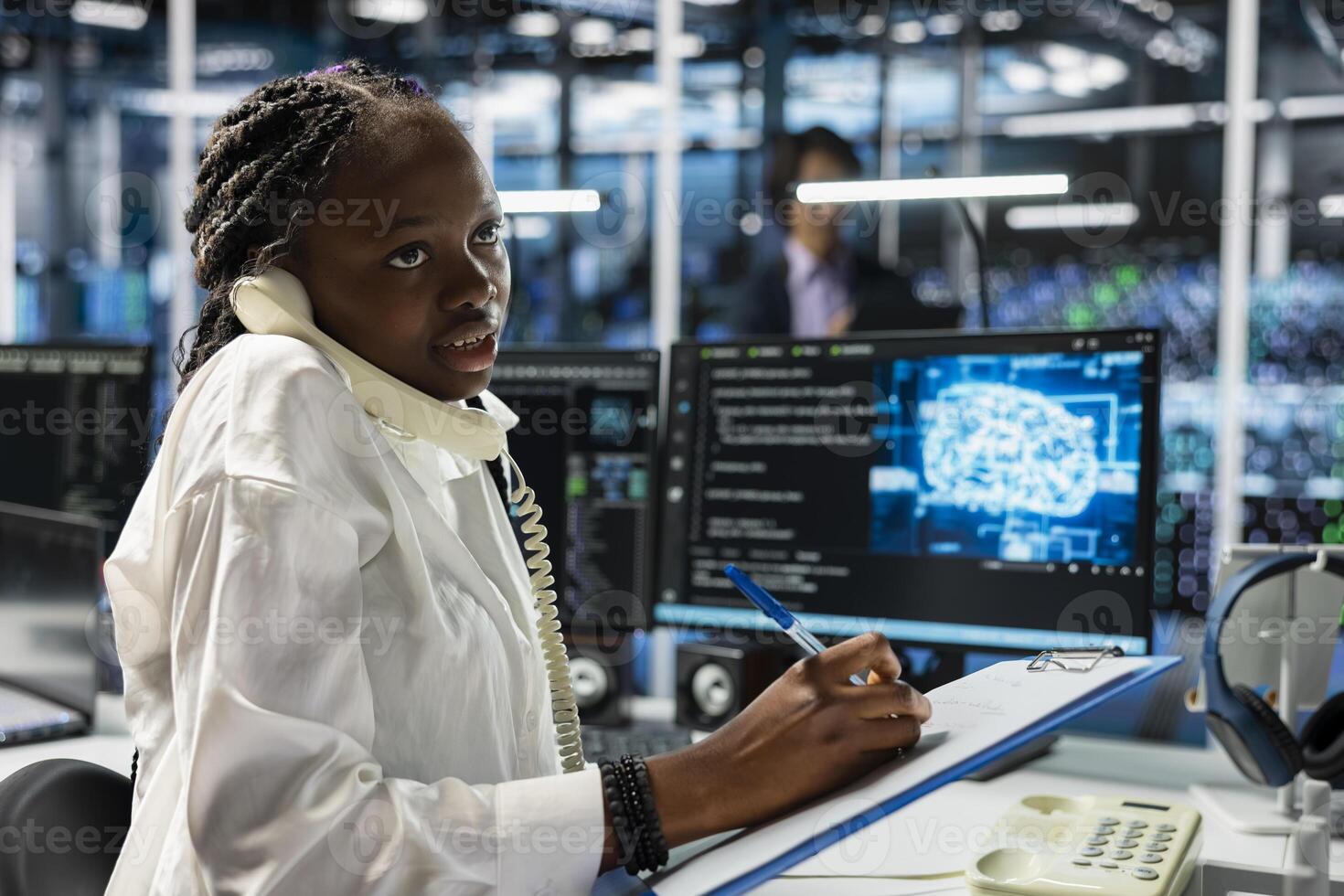 Data center operator answers telephone call, assisting users, taking notes. African american woman in server farm talks in phone call, managing clients issues using artificial intelligence photo