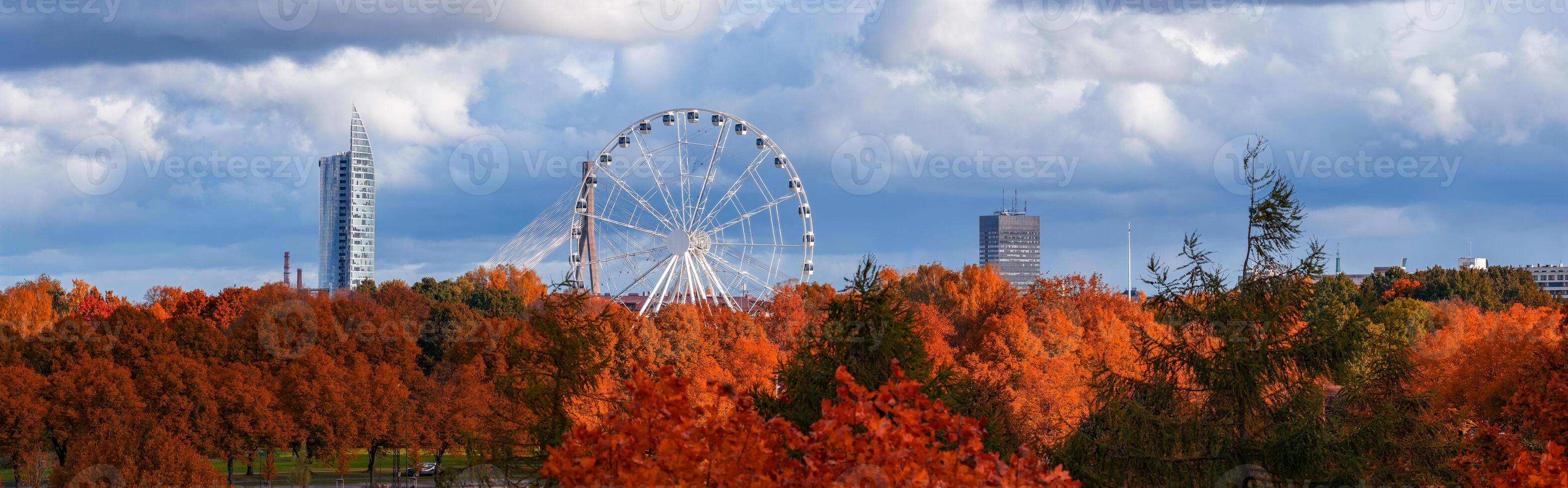 Ferris wheel rises over orange and red trees in Riga, Latvia, with high rises and a triangular tower. Dramatic clouds and crisp daylight highlight peak fall color. photo