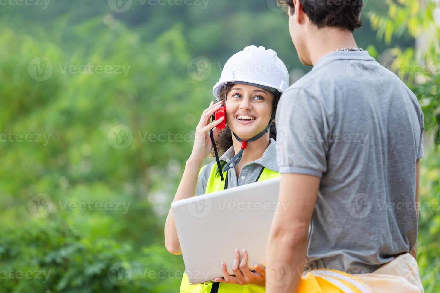 Smiling Female Engineer Communicating with Walkie Talkie on Site, Construction Manager Using Laptop and Radio for Team Coordination, Happy Project Supervisor Discussing Work Outdoors with Colleague photo