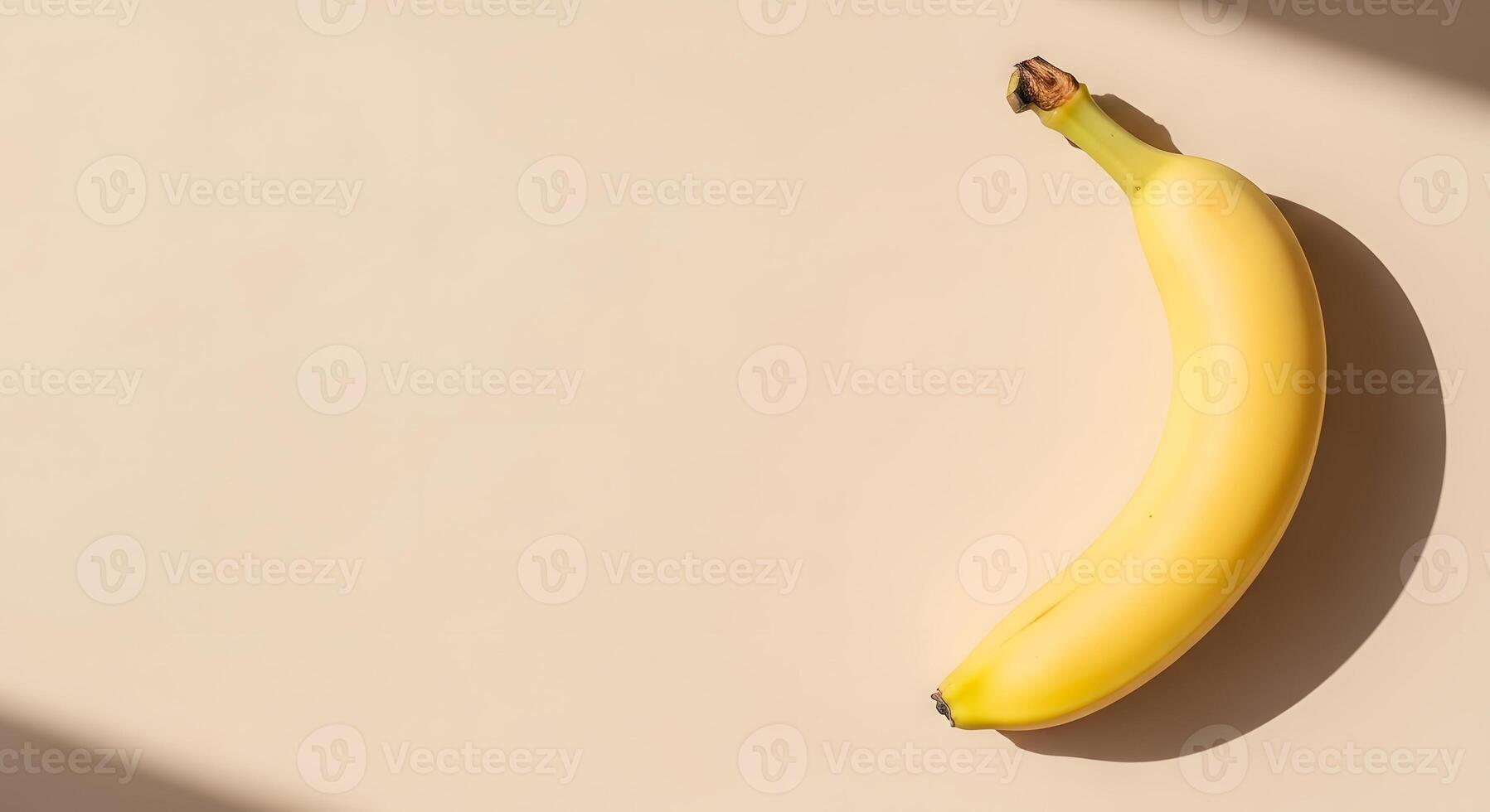 A single, bright yellow banana casts a long shadow on a plain, light-colored background, highlighting its simple shape and fresh appearance photo