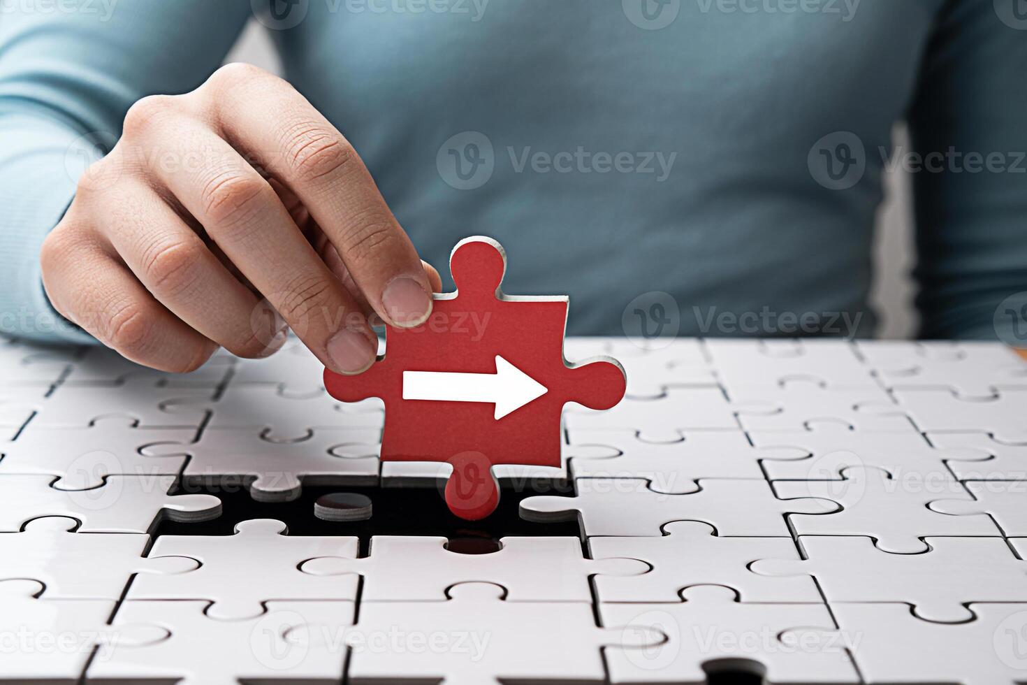A hand carefully placing a red puzzle piece with a white arrow into a jigsaw puzzle on a white table symbolizing progress direction and strategic problemsolving with a focus on achieving business goal photo