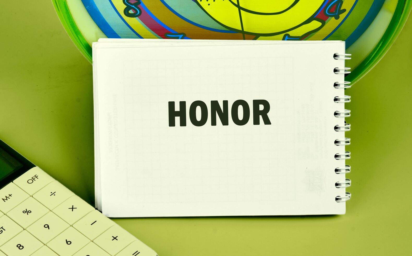 A spiral notebook opened to the word honor is placed alongside a calculator and a colorful clock, creating a studious atmosphere perfect for learning and reflection photo