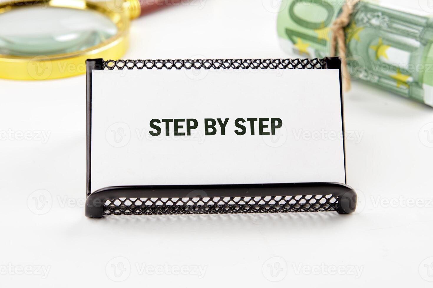 A small black stand holds a white card that reads STEP BY STEP, surrounded by a magnifying glass and rolled currency. The setting suggests a focused approach to tasks photo