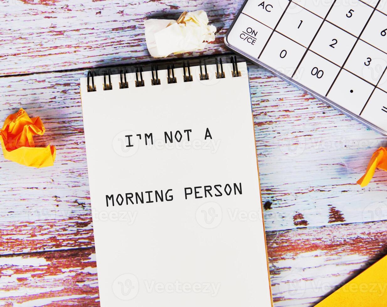 A notepad with the words I am not a morning person sits on a wooden table next to a calculator and crumpled papers, reflecting the challenge of starting the day early and creatively photo