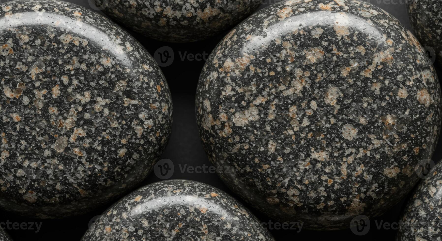 Close-up showcases smooth, round dark granite stones with speckled beige and gray inclusions on a matte black surface, creating a natural pattern. photo
