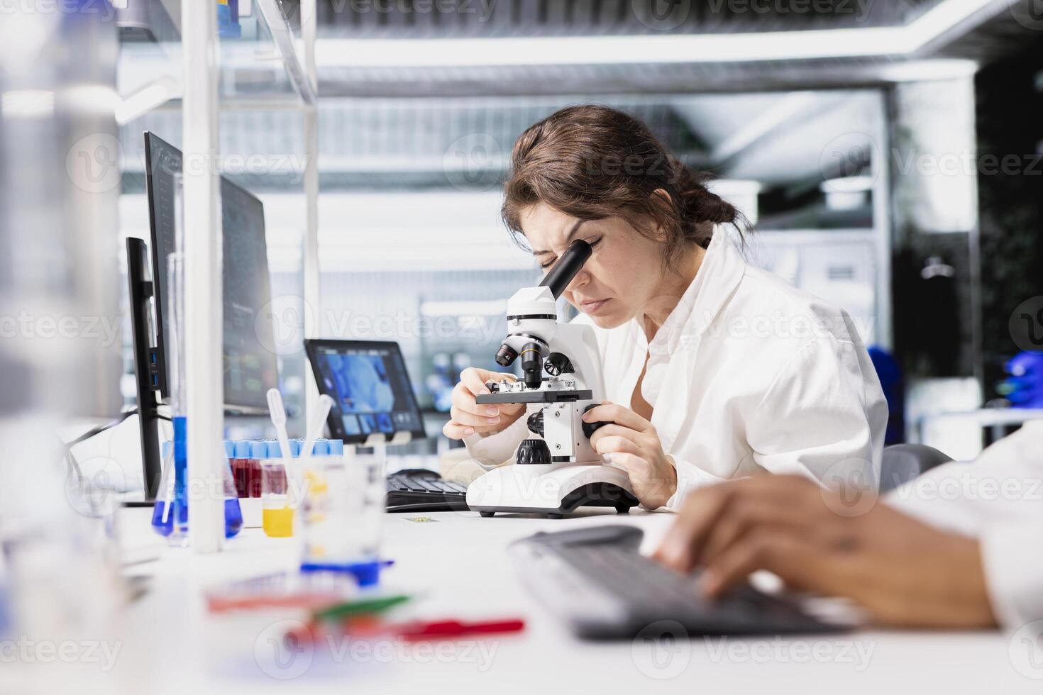 Cytotechnologist adjusting microscope, zooming on cellular structures in lab workspace. Molecular biologist observes specimens through magnification tool at laboratory bench workstation photo