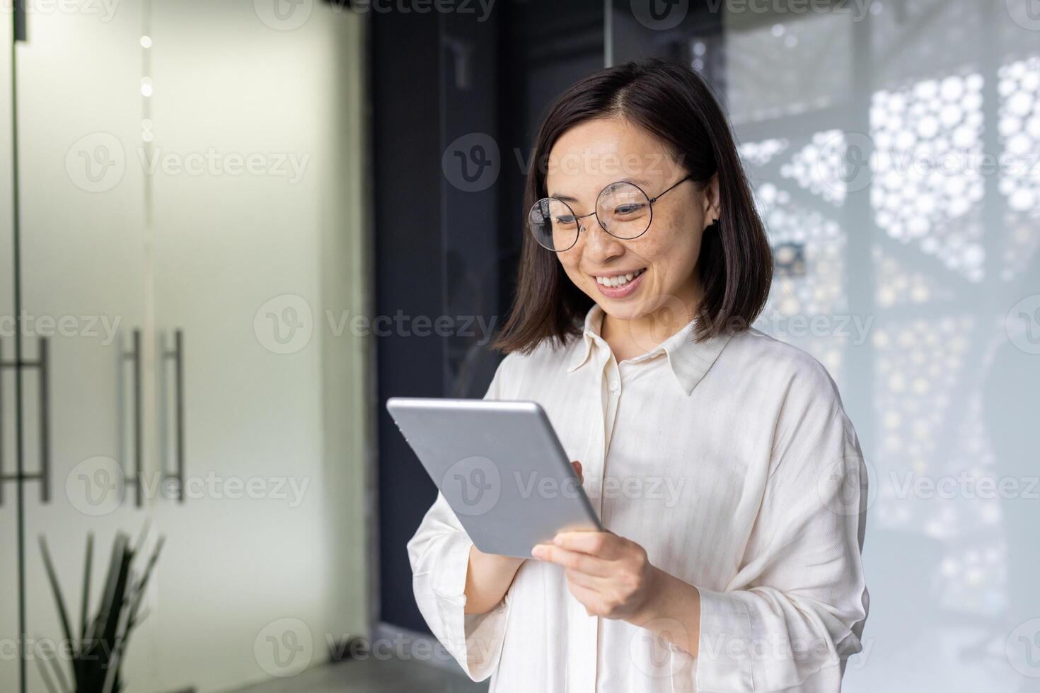 Young beautiful Asian woman with tablet computer standing near window, smiling businesswoman using online application, programmer testing new software. photo
