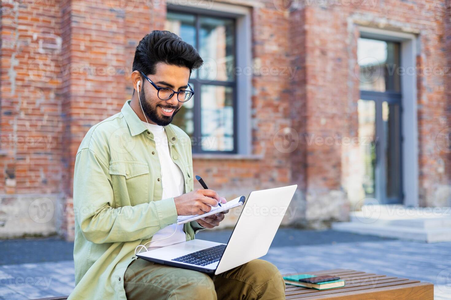 Young man studying remotely sitting on a bench outside a university building, a student in headphones watching a online course, talking using a call, writing down data in a notebook. photo