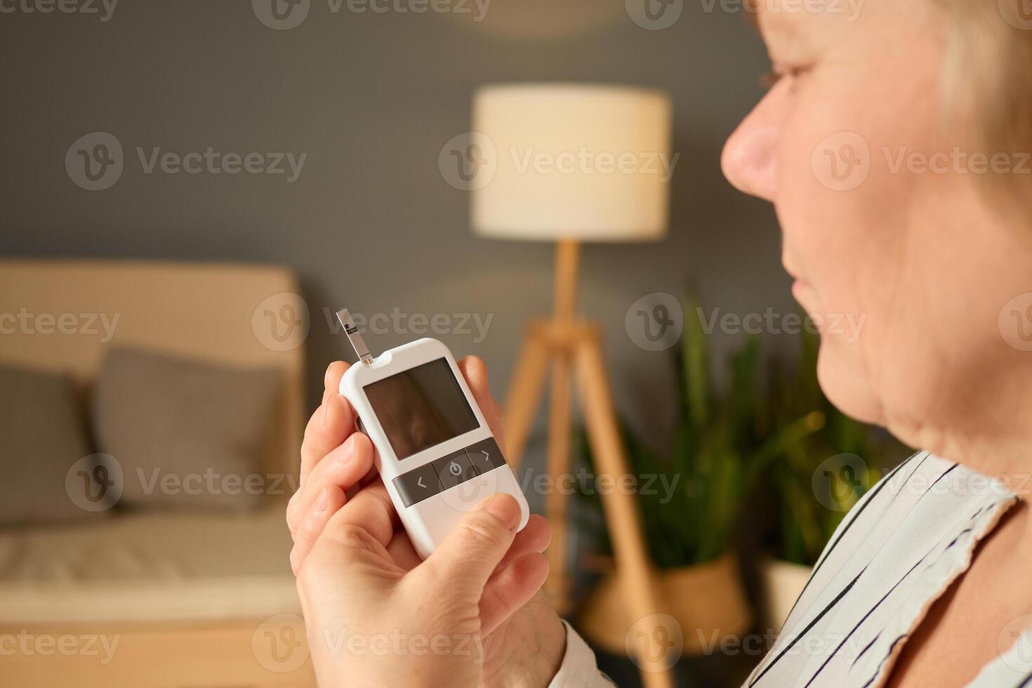 A person checks their blood sugar levels using a glucometer while seated in a comfortable living room. The atmosphere is warm and inviting, with a lamp and plants in the background photo
