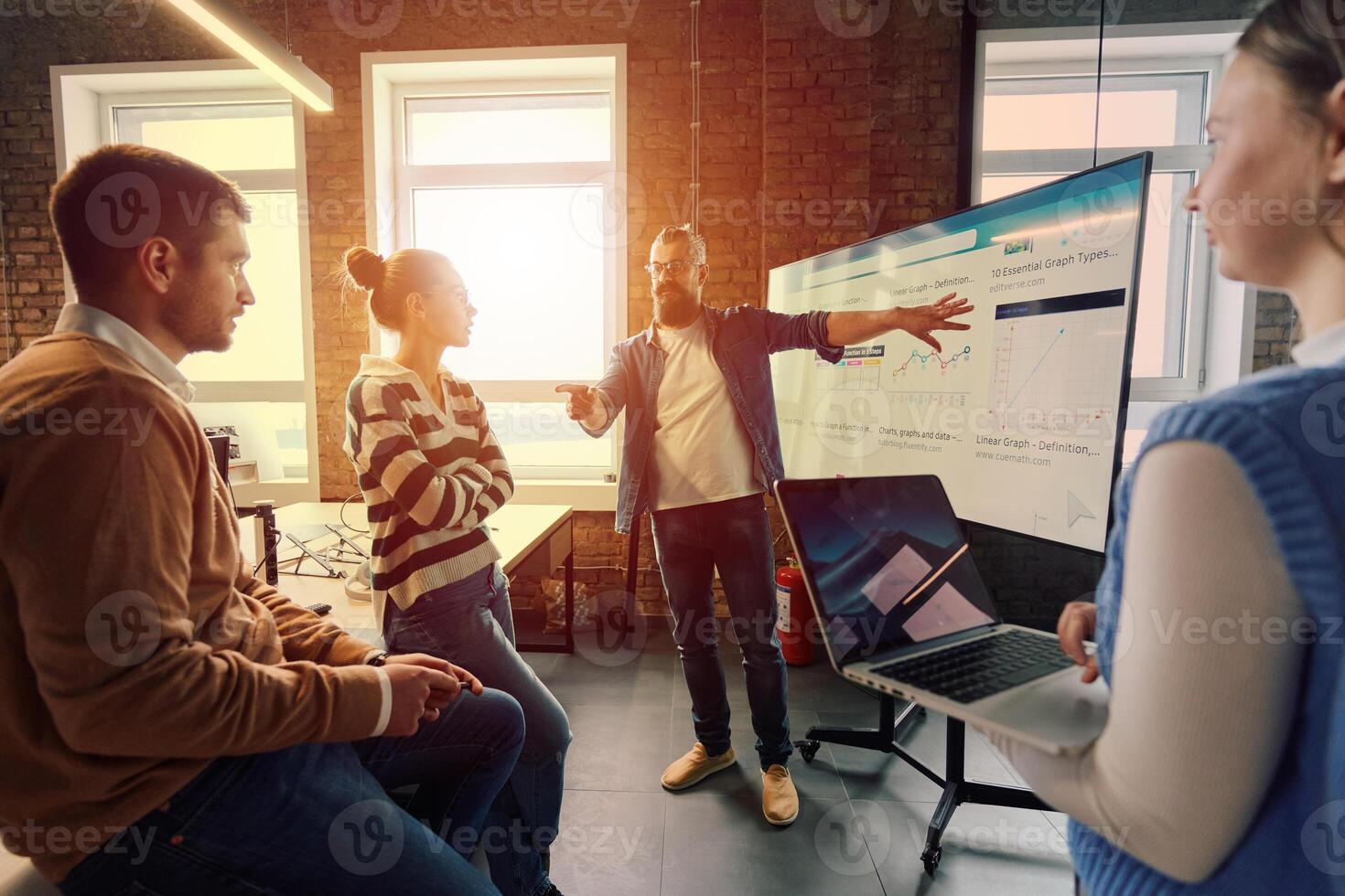 Group of professionals gathered in a modern office for a business presentation. A man with a beard explains data charts on a large screen while the team listens attentively. Concept of teamwork photo