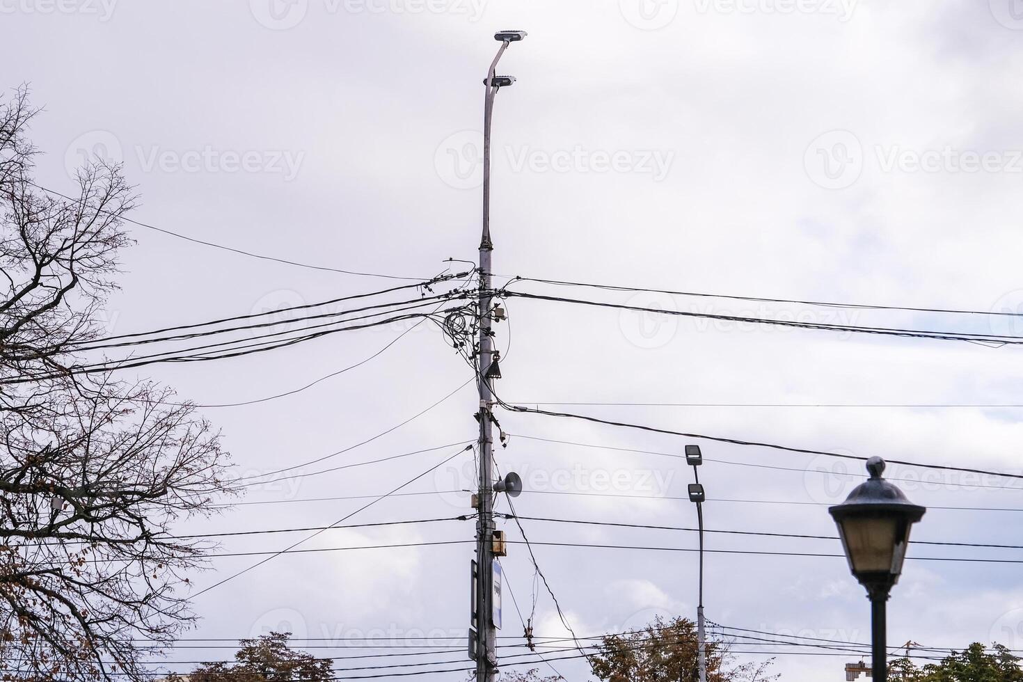 Tangled power lines and light poles against a cloudy grey sky with bare tree branches, showing urban infrastructure and electrical grid chaos. photo