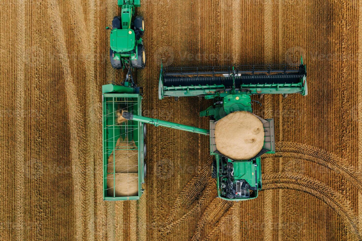 An aerial view shows a green combine harvester unloading grain into a green tractor with a trailer on a golden brown field with visible tire tracks. photo