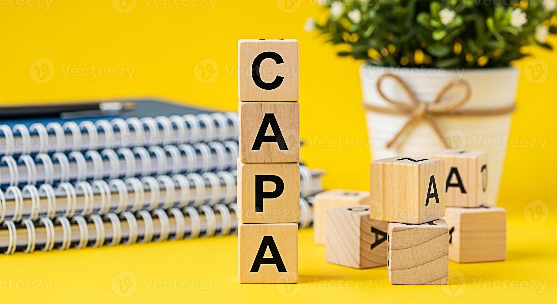Wooden blocks spelling CAPA standing on a yellow desk with notebooks and a potted plant representing corrective action and preventative action for quality control and continuous improvement photo