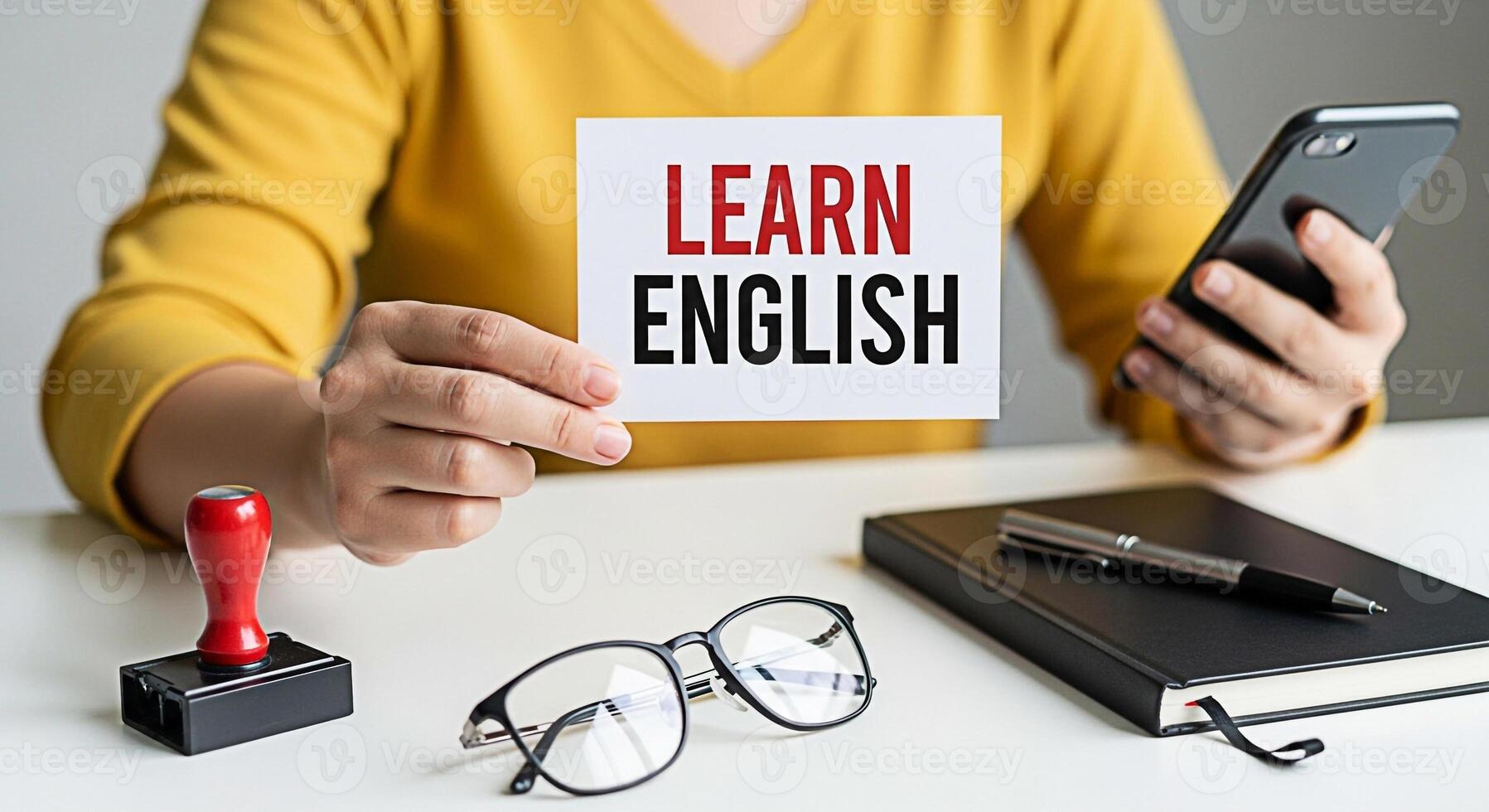 Student holding a Learn English sign while studying with a smartphone notebook pen glasses and stamp on a white desk representing education language learning and a positive motivated learning environm photo