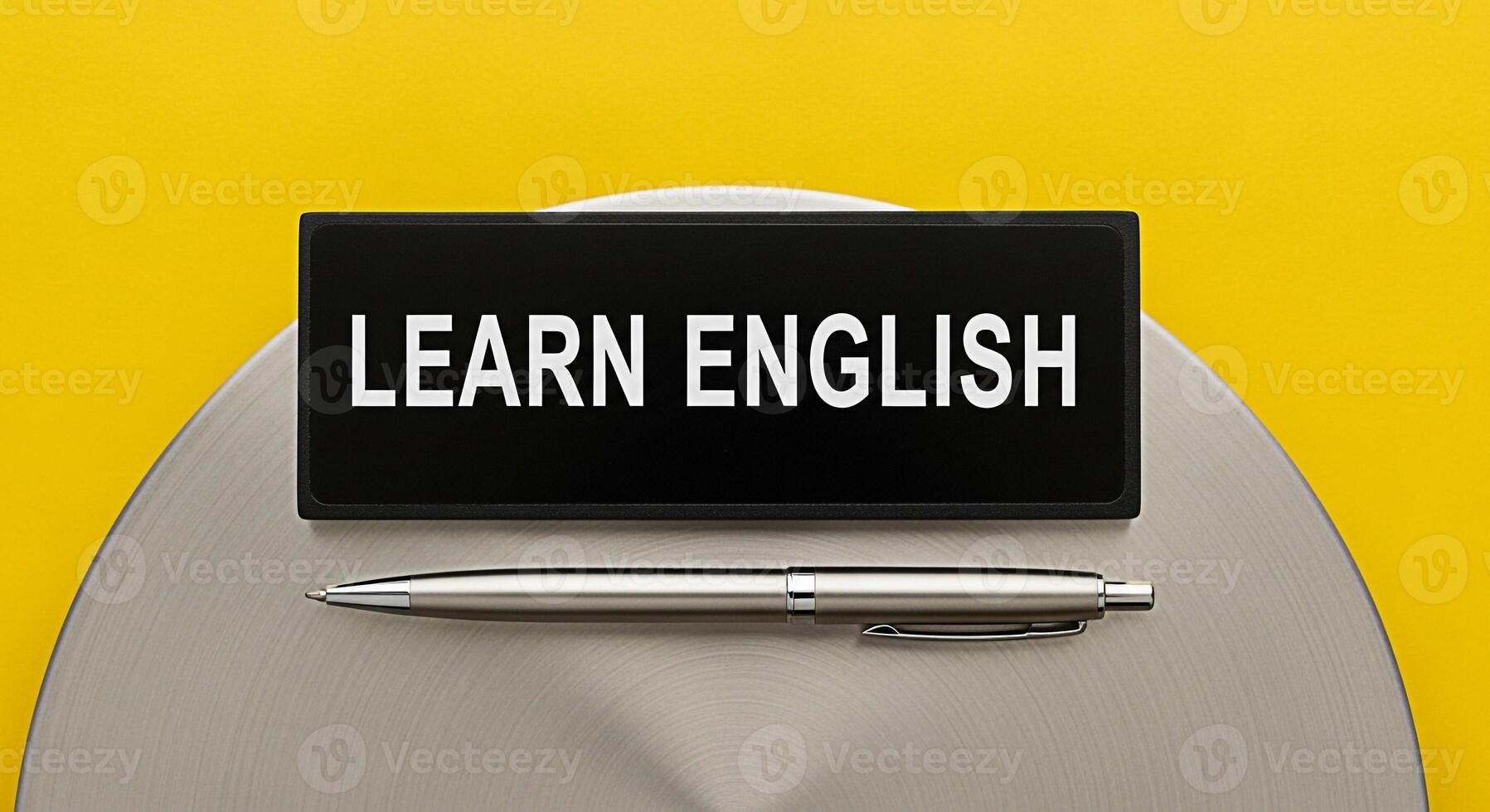 Closeup of a black sign displaying Learn English with a sleek silver pen resting on a metallic surface against a vibrant yellow background symbolizing education and the pursuit of language skills photo