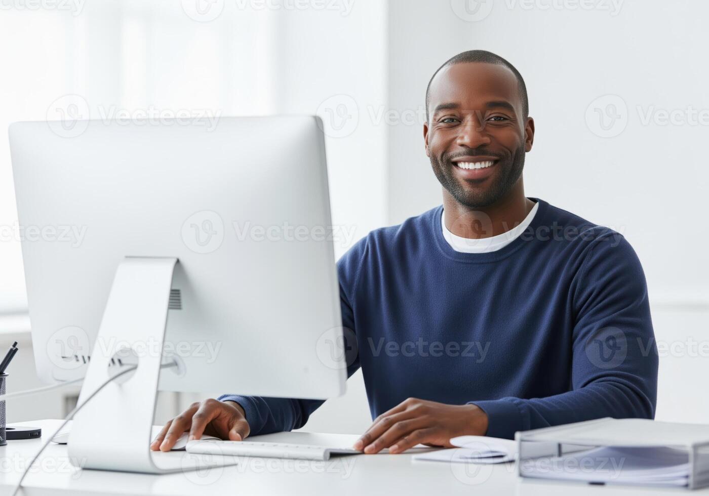 Confident black man smiling while working on a computer at a modern white office desk photo