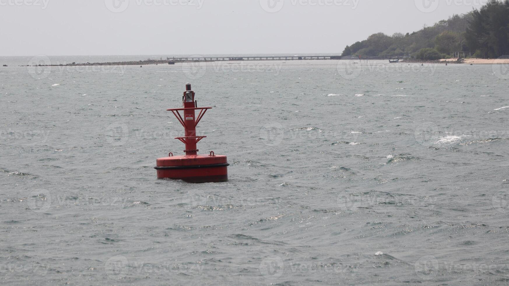 Red buoy floats in the ocean near a distant pier and coastline on a cloudy day, creating a sense of maritime navigation and safety. photo