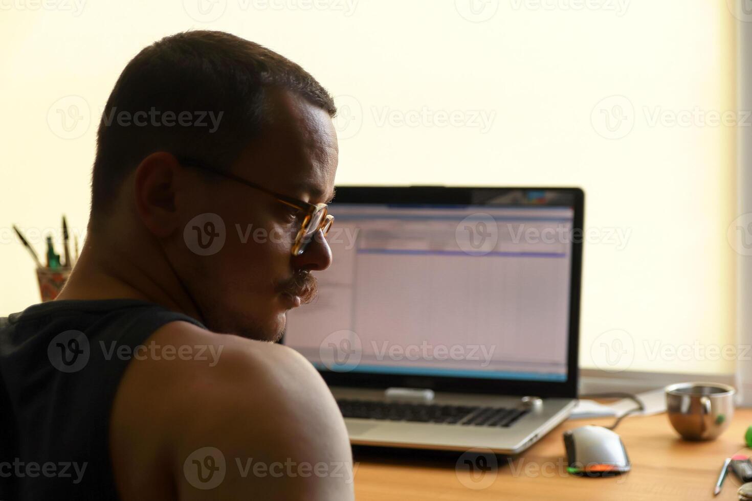 A portrait of a man in profile, seated with his laptop. His thoughtful expression, highlighted by his eyewear and moustache, hints at procrastination or deep, focused thought while working photo