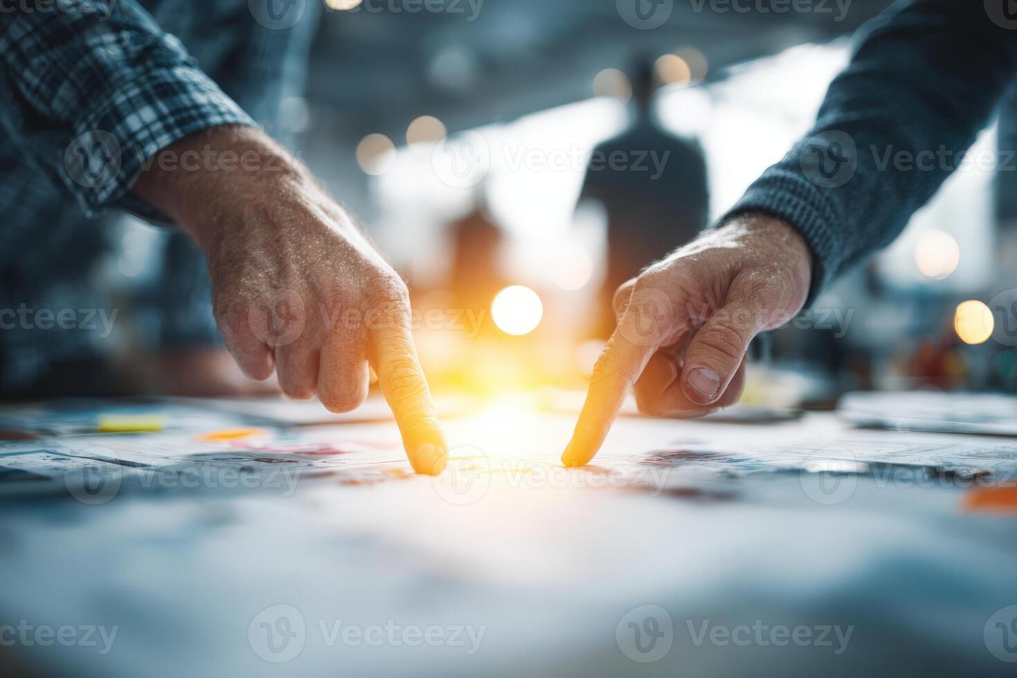Two hands pointing at a document on a table, illuminated by warm light, surrounded by blurred figures in the background, showcasing collaboration and teamwork in a creative workspace photo
